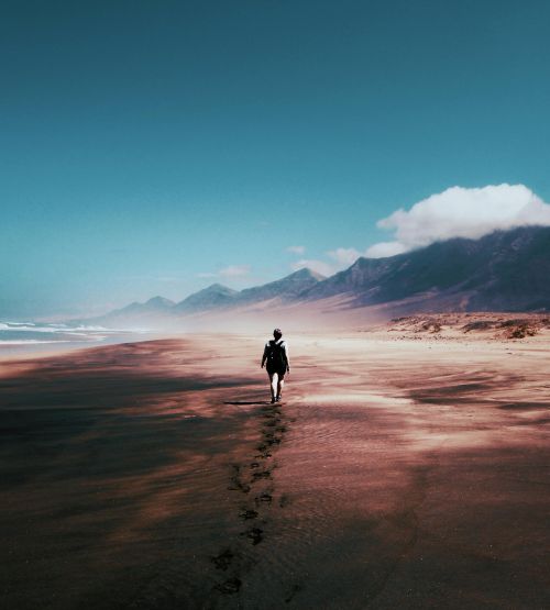 A lone person walking along a vast sandy beach in Pájara, Spain, with mountains in the background.
