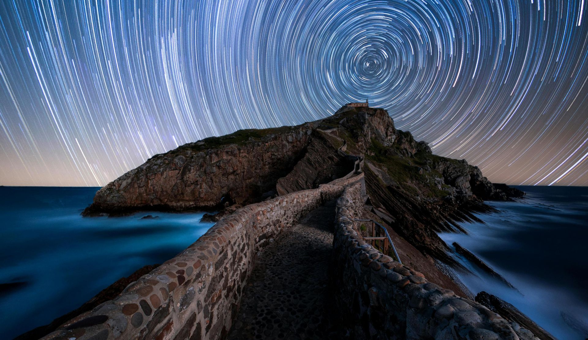 Stunning star trails over a rocky island with a winding stone pathway at night.